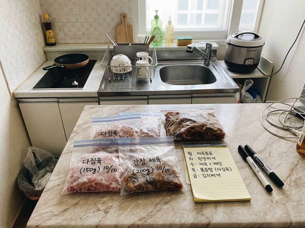 조리대 위 다짐육·간장 제육 지퍼백들과 요일별 메뉴 메모, 펜 두 자루